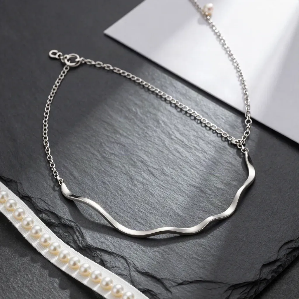 Zilveren golvende ketting met ringetje en parelwit textiel op matte leisteen.